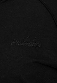 Zwarte stof met subtiel zwart geborduurd woord „smilodox“ in cursief op het oppervlak.