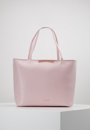 Sac cabas en cuir rose clair avec doubles poignées et petit détail nœud doré sur une surface blanche unie.