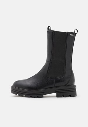 Botte mi-mollet en cuir noir avec panneaux latéraux élastiques, languettes de tirage, semelle texturée et talon bas, présentée sur un fond gris clair.