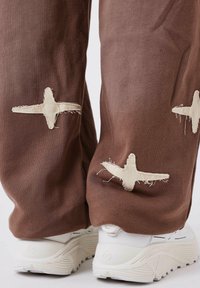 Pantaloni di tessuto marrone con ricami crema a forma di stella; abbinati a sneaker bianche con suole testurizzate e dettagli sottili.