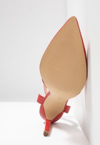 Chaussure rouge à talon haut avec bout pointu, semelle orientée vers l'avant montrant une adhérence texturée et la taille 37, posée contre des murs blancs.