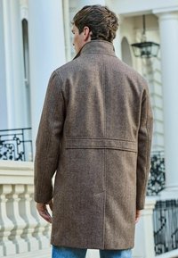 Ruskea herringbone-villakangastakki, jossa on korkea kaulus, taka-aukko ja rento istuvuus, yhdistettynä vaaleansinisiin farkkuihin.