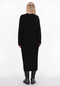Maglione lungo nero lavorato a maglia con texture a coste e scollatura rotonda, dotato di maniche lunghe e taglio dritto. Indossato con stivali neri con fibbia.