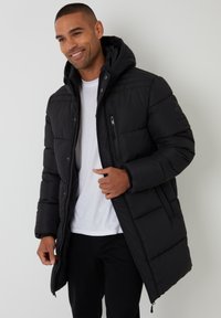 Black puffer jacket with a hood, featuring a zippered pocket, quilted pattern, and elastic cuffs, worn over a white t-shirt and dark pants.