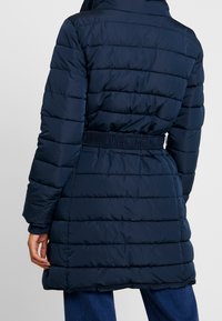 Chaqueta acolchada azul marino con cintura ajustada, que presenta una textura acolchada, mangas largas y puños de canalé. La vista trasera muestra la capucha.