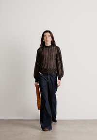 Blusa nera, con texture, a maniche lunghe e a palloncino, con una vita arricciata; abbinata a jeans ampi in denim scuro; la modella porta una cintura a tracolla beige.