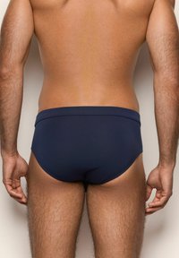 Hombre adulto vestido con calzoncillos azul marino, mostrado desde atrás, de pie con los brazos relajados a los lados del cuerpo frente a un fondo sencillo.