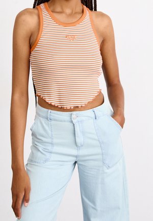 Frau trägt ärmelloses, orange-weiß gestreiftes Crop-Top und hellblaue hoch taillierte Hose mit einer Hand in der Tasche vor weißem Hintergrund.
