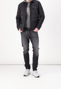 Schwarze gesteppte Jacke mit Druckknöpfen und Brusttasche, getragen über einem grauen Hoodie. Dunkle Jeans und weiße Sneakers runden das Outfit ab.