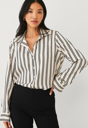 Woman wearing a beige button-up shirt with black vertical stripes and black high-waisted pants, posing with one hand on her hip.