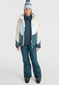 Veste de ski crème, bleu clair et vert foncé avec fermeture éclair ; associée à un pantalon teal et des bottes de ski noires ; comprend un bonnet bleu avec logo.