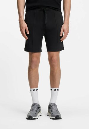 Άνδρας που φοράει μαύρα σορτς με κορδόνι, λευκές κάλτσες με μαύρο κείμενο και γκρι παπούτσια τρεξίματος, στέκεται σε λευκό φόντο.