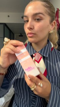 Młoda kobieta w pasiastym szaliku i wzorzystym szaliku trzyma pudełko z produktem do pielęgnacji skóry oznaczone "cześć wspaniała" w pomieszczeniu.