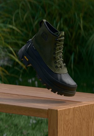 Зелен и черен висок водоустойчив ботуш с дебела подметка Vibram, висящ над дървена пейка на открито с фон на трева.