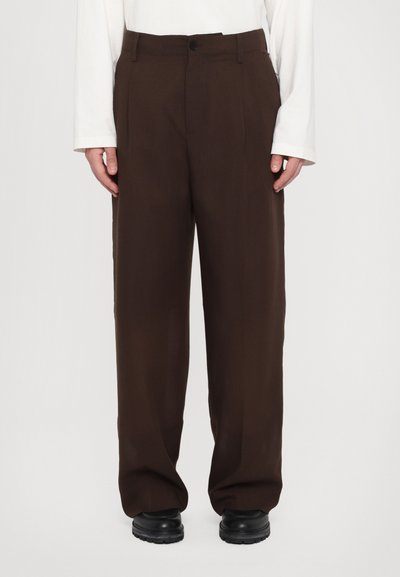 Brown tailored trousers with a high waist, pleats, and a straight leg. Made of smooth fabric, paired with black shoes.