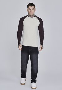 Raglanærmet skjorte i beige med mørkebrune ærmer, kombineret med mørke jeans og hvide sneakers. Har en rund halsudskæring og logodetalje.