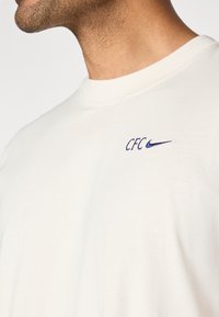 Jasnokremowa bawełniana koszulka z okrągłym dekoltem i krótkimi rękawami. Na piersi znajduje się haftowane logo "CFC" oraz niebieski znak Nike.