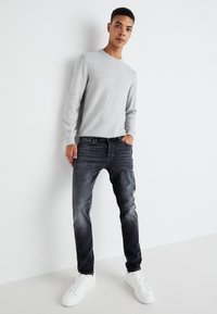 Pulover gri cu mâneci lungi, croială lejeră, asortat cu blugi skinny de culoare închisă și adidași albi. Fundalul este simplu și deschis.