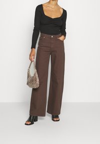 Donna che indossa un top fitted a maniche lunghe nero e pantaloni brown a vita alta e a gamba larga, mentre tiene in mano una borsa beige in pelle di serpente e una paio di scarpe con tacco aperto nere.