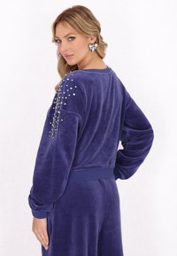 Lila velourtröja med långa ärmar, prydd med silverfärgade nitar på vänster axel. Ribbad mudd och midja. Mjuk textur.