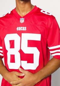 Camiseta roja de la NFL con el número "85" en blanco y el texto "49ERS". Cuenta con mangas a rayas y un cuello redondo. Hecha de un tejido ligero.
