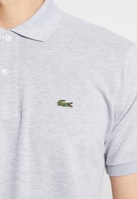 Polo de color gris claro con mangas cortas y dos botones en la parte delantera, que presenta un pequeño logo de cocodrilo verde en el lado izquierdo del pecho.