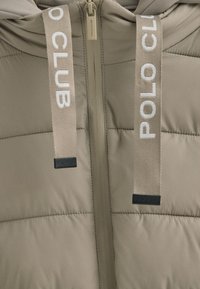 Giubbotto puffer di un beige chiaro con cuciture orizzontali, dotato di chiusura a zip e logo "POLO CLUB" su fasce in tessuto. Texture liscia.
