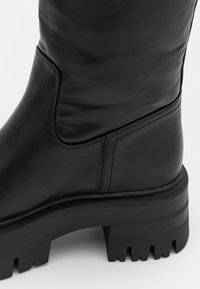 Stivaletto in pelle nera con design alto, tomaia testurizzata, cuciture rinforzate e suola in gomma spessa e robusta con dettagli di trazione.