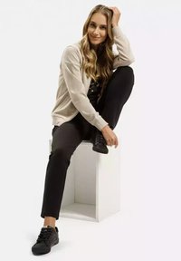 Beżowa kurtka zapinana na zamek, czarny top, czarne spodnie i czarne sneakersy. Modelka siedząca na białym sześcianie z długimi włosami ułożonymi w fale.