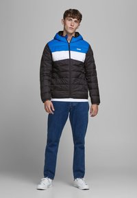 Gevoerde jas met een blauw en witte bovenkant en een zwarte onderkant. Heeft een capuchon, een volledige ritssluiting en wordt gedragen over een wit shirt en jeans.