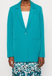 Tyrkysové sako s jedním knoflíkem, klopami s výstřihem do „V“ a dvěma předními kapsami. Vyrobeno z hladké tkaniny, v kombinaci s květinovou sukní.