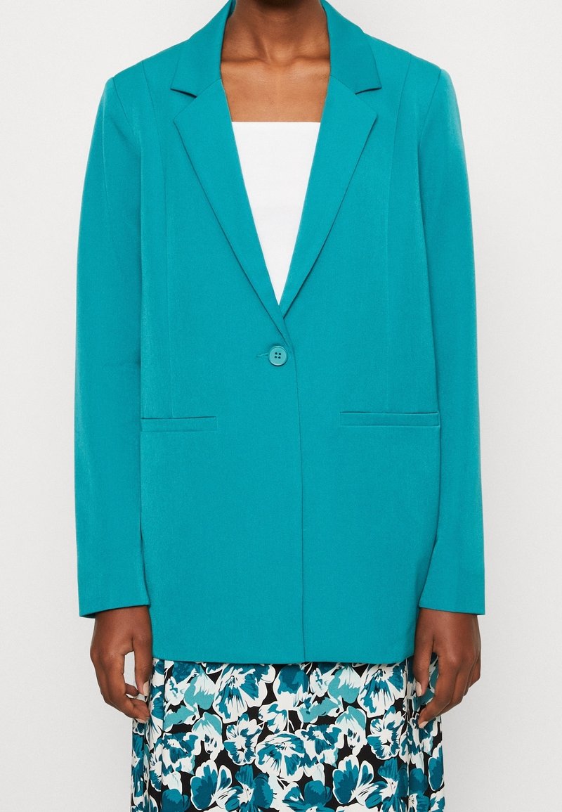 Tyrkysové sako s jedním knoflíkem, klopami s výstřihem do „V“ a dvěma předními kapsami. Vyrobeno z hladké tkaniny, v kombinaci s květinovou sukní.