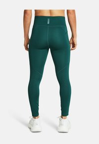 Osoba w ciemnozielonych legginsach o wysokim stanie z perforowanymi detalami na kolanach i białych butach sportowych, widziana z tyłu.