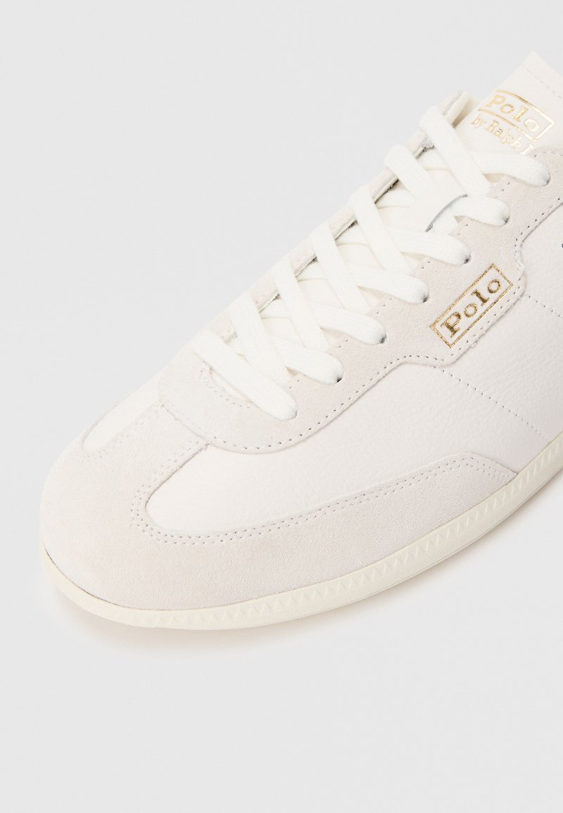 Scarpa da ginnastica bianca con tomaia in pelle e camoscio, lacci piatti, suola in gomma e dettaglio con logo dorato sul lato. Presenta rifiniture testurizzate.