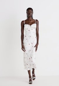 Abito midi bianco a fiori con silhouette aderente, busto arricciato, spalline sottili e delicati motivi a rosa. Indossato con sandali bianchi con lacci.