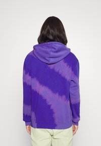 Fialová mikina s tie-dye vzorem, který postupně zesvětluje k fialové barvě, s kapucí na šňůrku a elastickými manžetami, vyrobená z jemné bavlněné látky.
