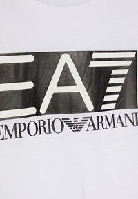 Bílý tričko s velkým černým grafikou a nápisem "EA7 EMPORIO ARMANI" a stylizovaným logem orla. Hladká textura, moderní design.