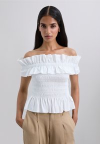 Kvinna iklädd en vit avskuren topp med volanger och beige byxor, stående med händerna i fickorna mot en ljus, enfärgad bakgrund.
