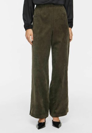 Pantalon en daim vert olive taille haute à jambes larges, porté avec un haut noir à manches longues et des talons noirs à bout pointu, debout sur un sol blanc.