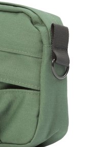 Grüne Stofftasche mit strukturierter Oberfläche, ausgestattet mit einem schwarzen Tragegurt und einem Metallring zum Befestigen sowie einem detailliere Fronttasche.