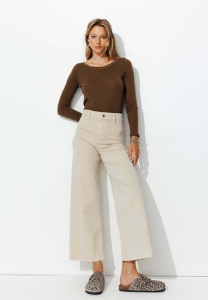 Haut marron à manches longues, pantalon beige taille haute à jambes larges avec ourlet effiloché, et chaussures à enfiler imprimé animal, sur fond blanc.