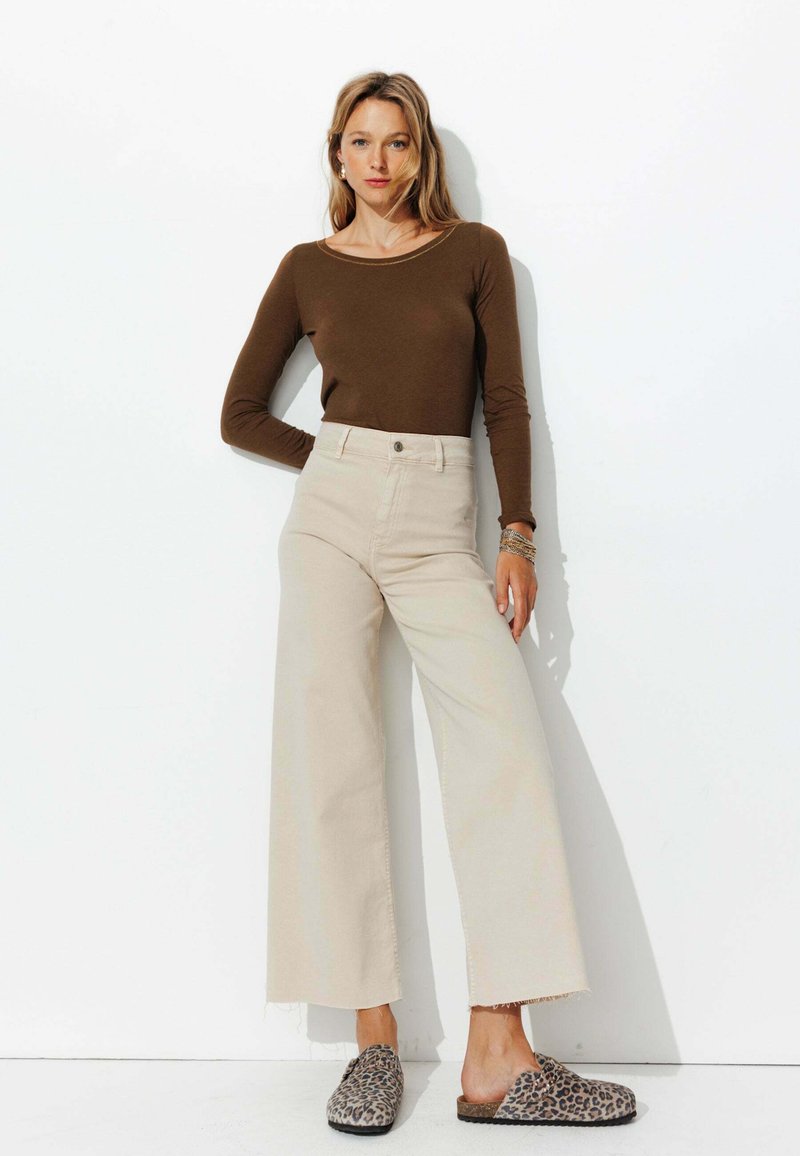 Haut marron à manches longues, pantalon beige taille haute à jambes larges avec ourlet effiloché, et chaussures à enfiler imprimé animal, sur fond blanc.