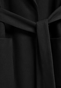 Tejido de abrigo negro con una textura suave. Presenta un cinturón atado en la cintura y dos bolsillos delanteros. El diseño es minimalista y moderno.