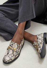 Osoba v šedých kalhotách a béžových mokasínech se vzorem hadí kůže a zlatou kovovou ozdobou, sedící na světlé podlaze.