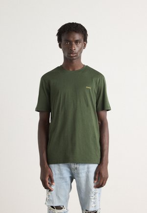 Grønn bomull T-skjorte med rund hals og korte ermer, med en liten gul "HUGO"-logo på venstre bryst. Brukt med slitte lyseblå jeans.