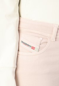 Jeans de mezclilla rosa claro con un bolsillo frontal que incluye una etiqueta, textura suave y detalles de costura sutiles. Parte de un atuendo casual.
