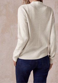 Osoba v krémovém hřejivém svetru a tmavě modrých džínách, zachycená zezadu na pozadí béžové texturované zdi.