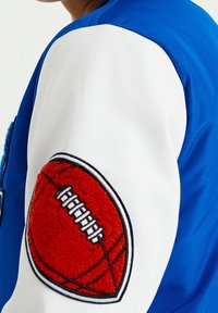 Modro-bílá bunda s texturovanou červenou fotbalovou nášivkou na rukávu. Bunda má hladký, lesklý povrch a kontrastní vzory.