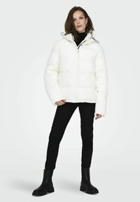 Hvid puffjakke med hætte, prydet med et quiltet design, trykknapper og elastiske manchetter. Sammen med sorte skinny jeans og støvler.