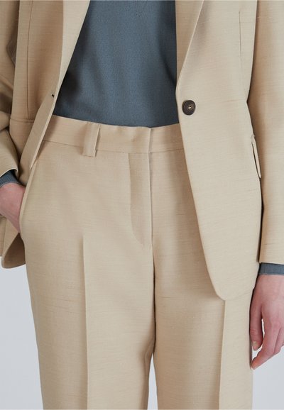 Beige op maat gemaakte broeken van gestructureerde stof, met een platte voorkant, zijzakken en een subtiele glans, gecombineerd met een grijze trui.
